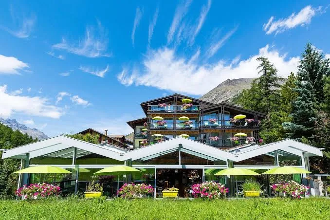 Wellness & Spa Hotel Pirmin Zurbriggen with glass ground floor, wooden upper floors, lime-green umbrellas and flower balconies against alpine mountains